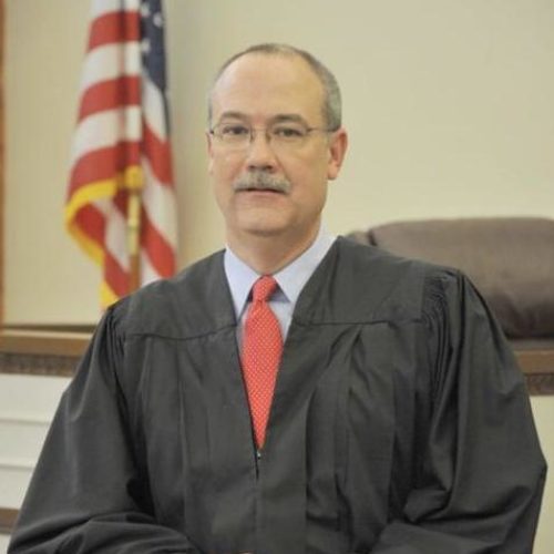 portrait of Judge Goss in a judge's robe sitting in front of an American flag