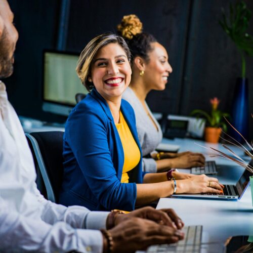 Woman in blue suit jacket smiles at her colleague while working on their laptops