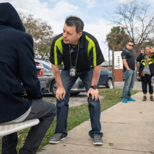 Police officer bends down to speak to man experiencing homelessness while two other officers wait to the side.