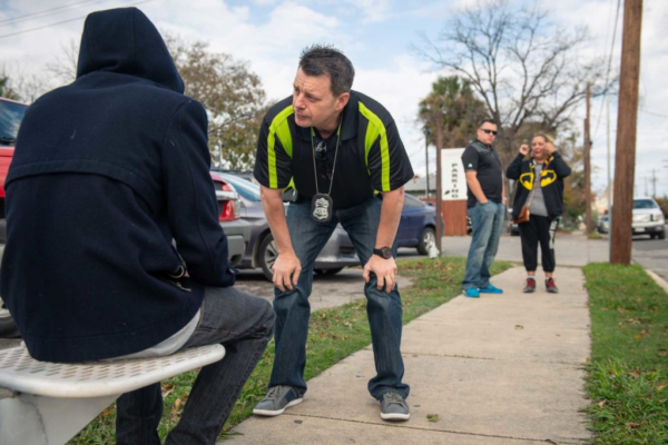 Police officer bends down to speak to man experiencing homelessness while two other officers wait to the side.