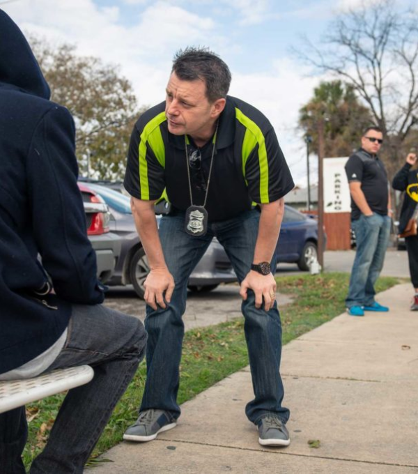 Police officer bends down to speak to man experiencing homelessness while two other officers wait to the side.