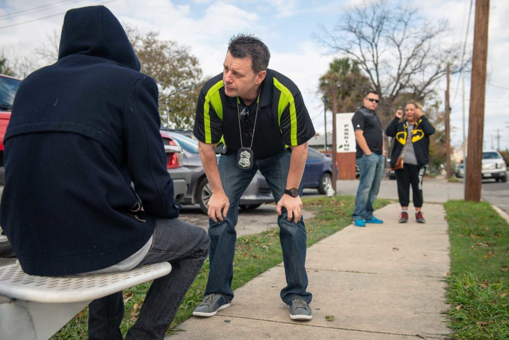 Police officer bends down to speak to man experiencing homelessness while two other officers wait to the side