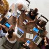 Eight people sitting around a table with laptops, notebooks, and phones. Two people are shaking hands.