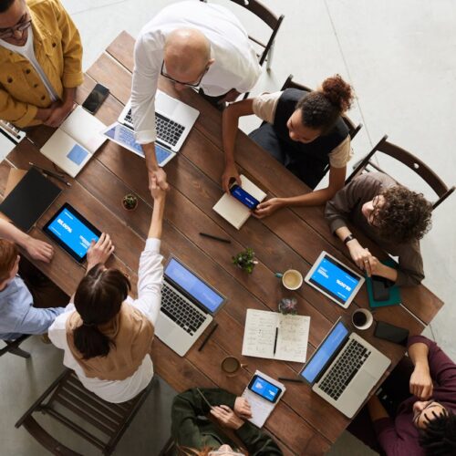 Eight people sitting around a table with laptops, notebooks, and phones. Two people are shaking hands.