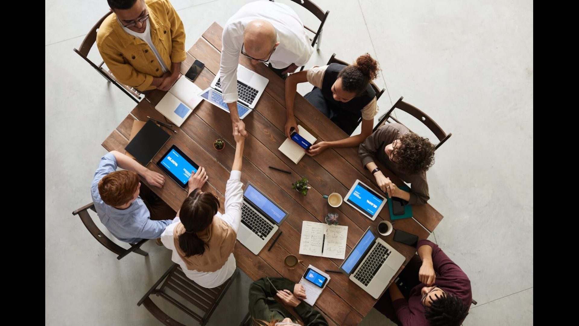 Eight people sitting around a table with laptops, notebooks, and phones. Two people are shaking hands.