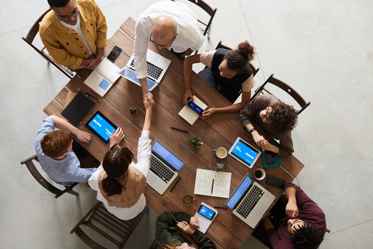 Eight people sitting around a table with laptops, notebooks, and phones. Two people are shaking hands.