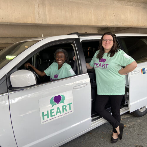 One woman sits in driver seat while another woman stands nearby a van with the Durham HEART logo on it. Both women have on shirts for Durham's HEART program.