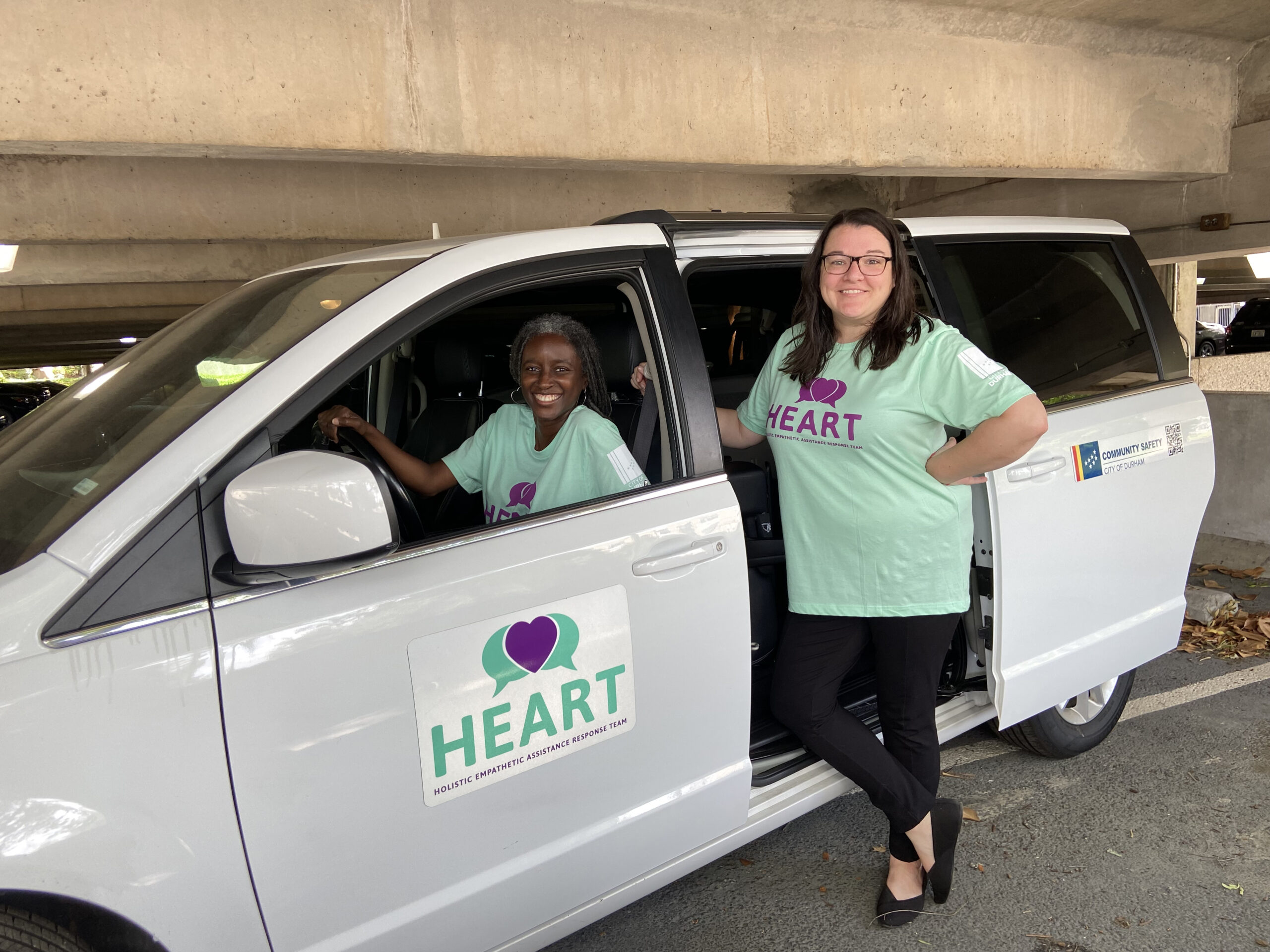 One woman sits in driver seat while another woman stands nearby a van with the Durham HEART logo on it. Both women have on shirts for Durham's HEART program.